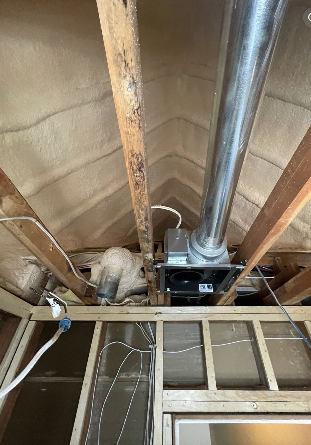 Spray foamed attic peak with a bathroom exhaust fan and duct being installed between rafters
