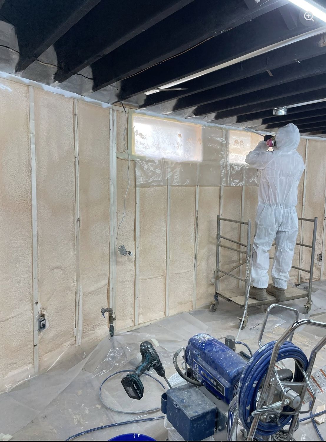 Installer in protective suit and respirator spray foaming a basement wall from a rolling scaffold, spray rig in the foreground