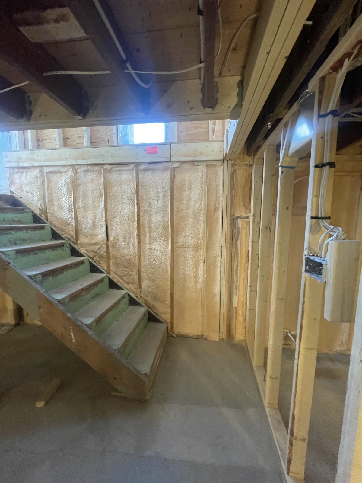 Basement stairwell wall spray foamed between studs with framing and electrical rough-ins visible