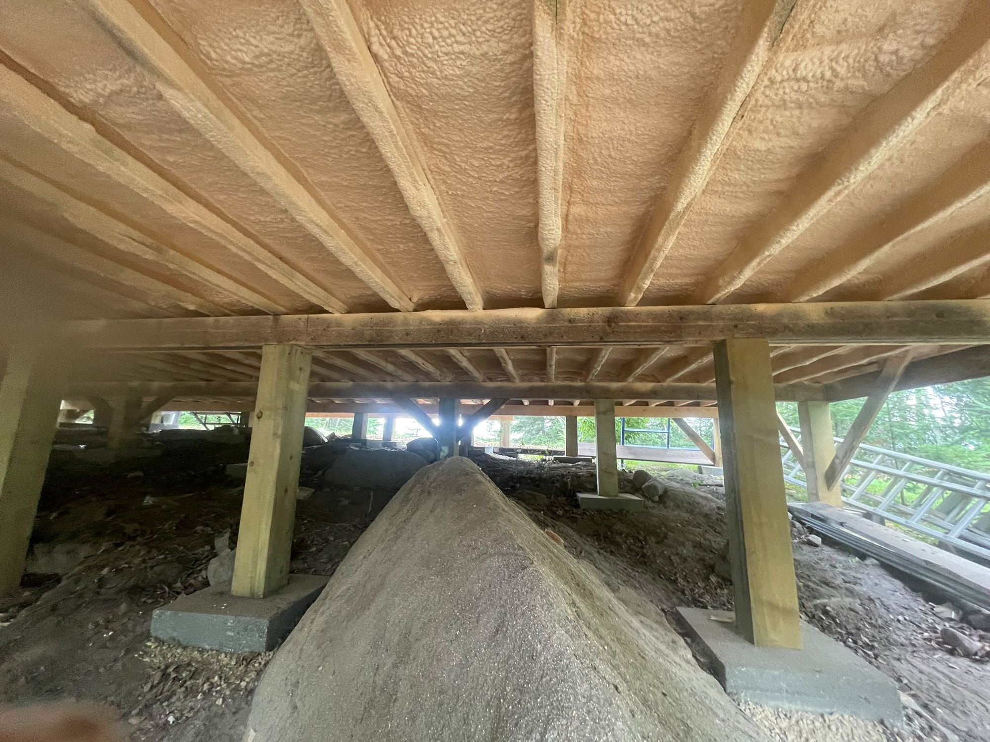 Underside of a cottage floor with closed-cell spray foam applied between the joists, built on a rock and post footing near the treeline