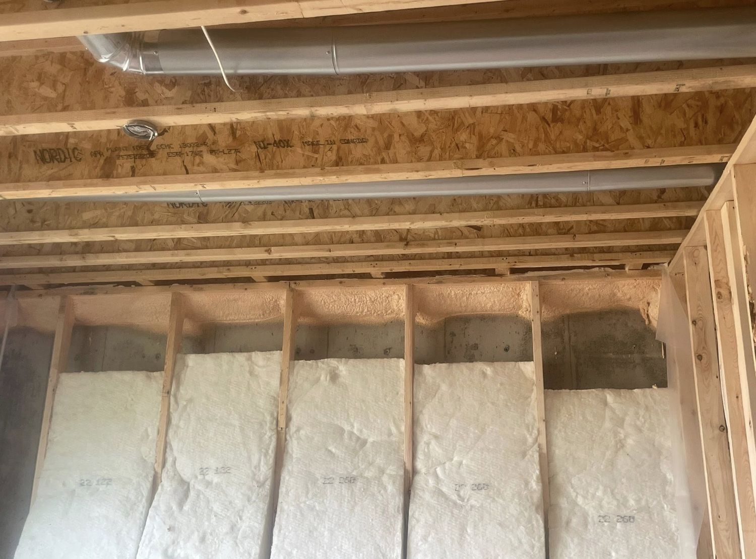 Spray foam sealing the rim joist above fiberglass batt insulation on a new basement wall, engineered floor joists overhead