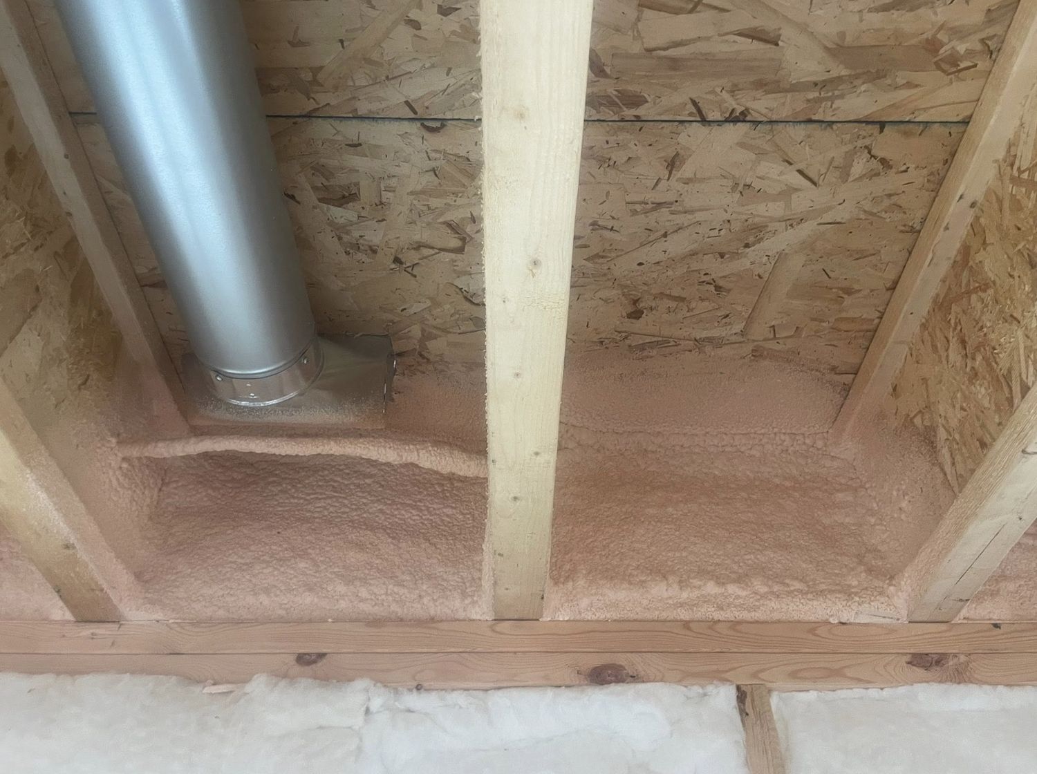 Close-up of spray foam sealing a rim joist cavity around a metal duct, batt insulation below
