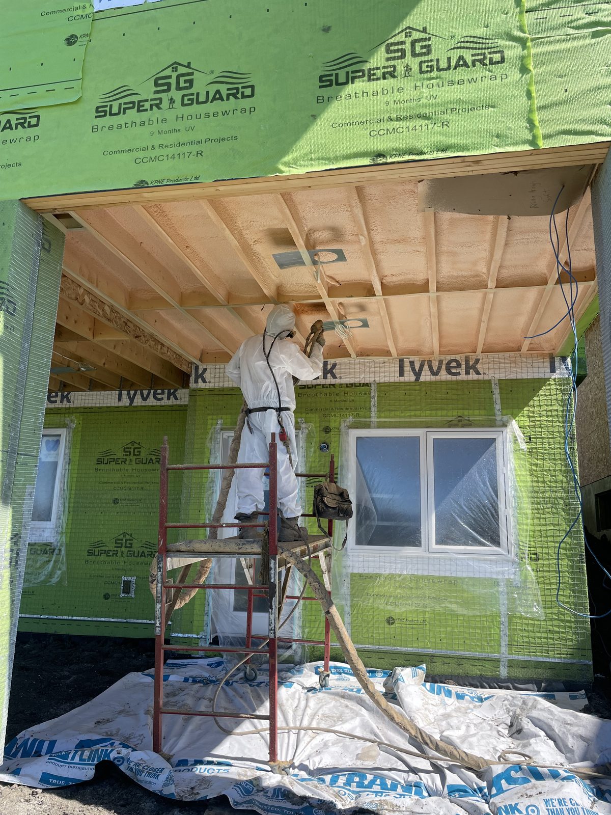 Installer in a hooded suit and full-face respirator spray foaming a covered entry ceiling from a rolling scaffold